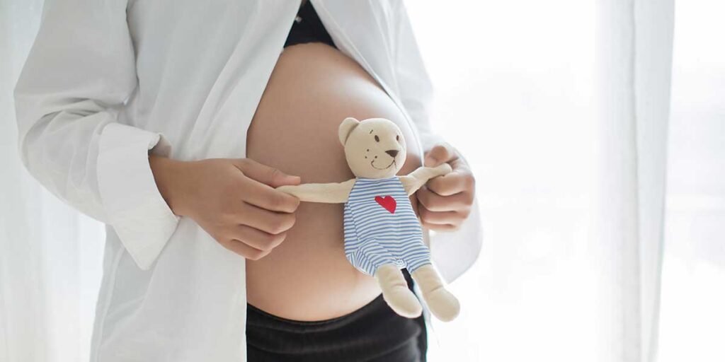 A pregnant woman gently holding a small stuffed teddy bear against her exposed belly, highlighting the importance of prenatal care.