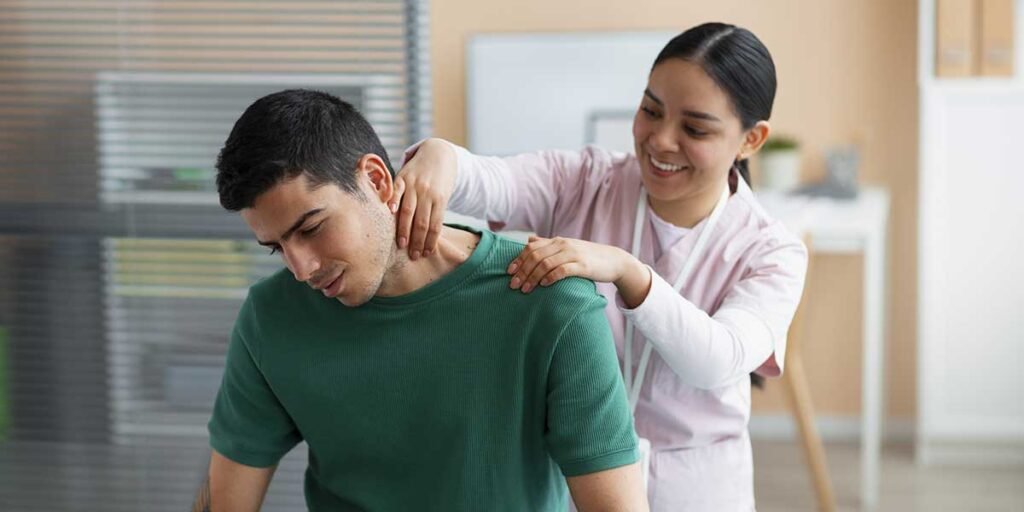 A smiling female chiropractor gently adjusting the shoulder and neck of a sitting male patient during a therapy session.