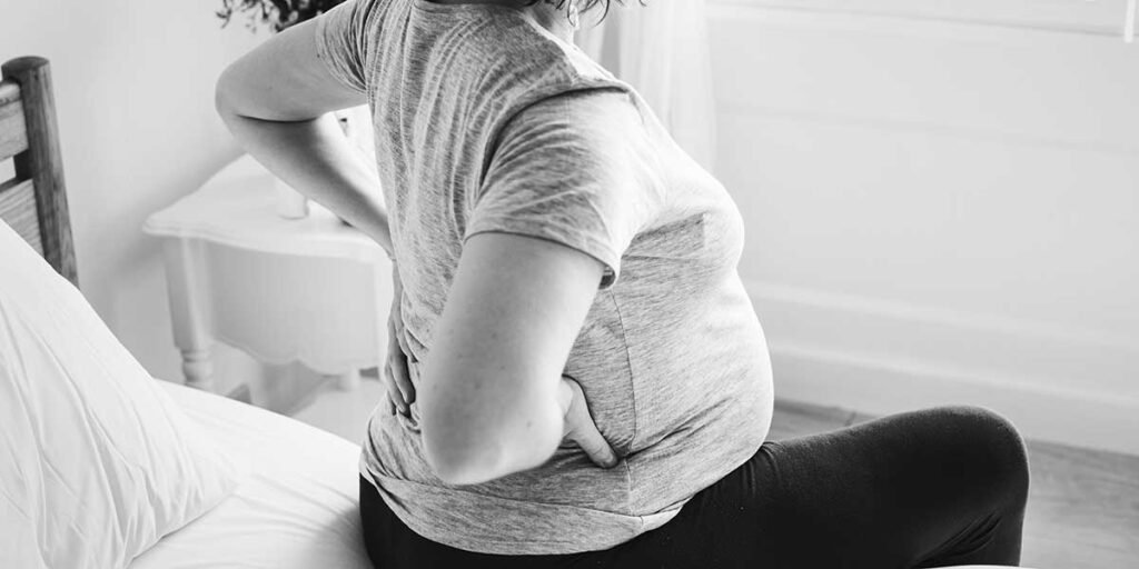 Black and white photo of a pregnant woman sitting on a bed while holding her lower back in discomfort.