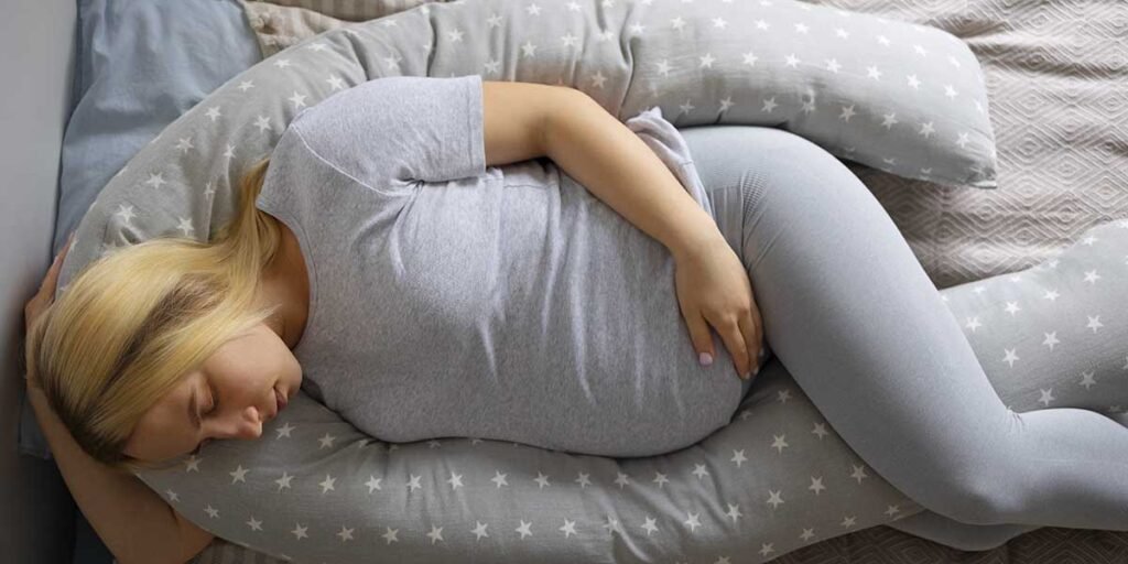 Pregnant woman sleeping comfortably on her side supported by a large grey star-patterned body pillow on a bed.