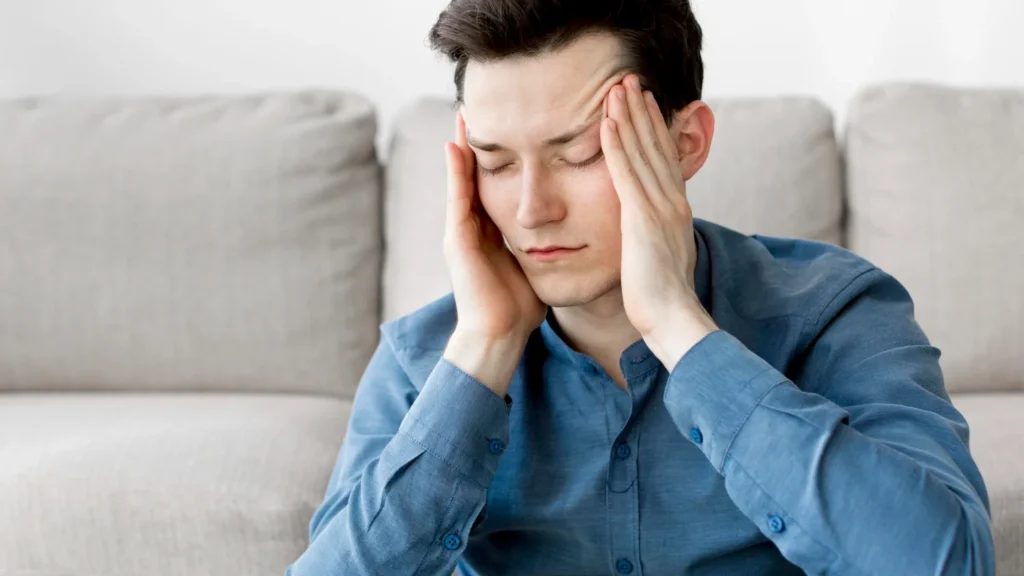 Man sitting on the floor touching his temples with eyes closed in discomfort due to severe migraines and headaches.